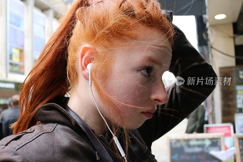 这是一个14 / 15岁的红发少女，皮肤苍白，脸上有雀斑，站在一个商店的橱窗里看着自己，一边用耳机听音乐，侧视图照片